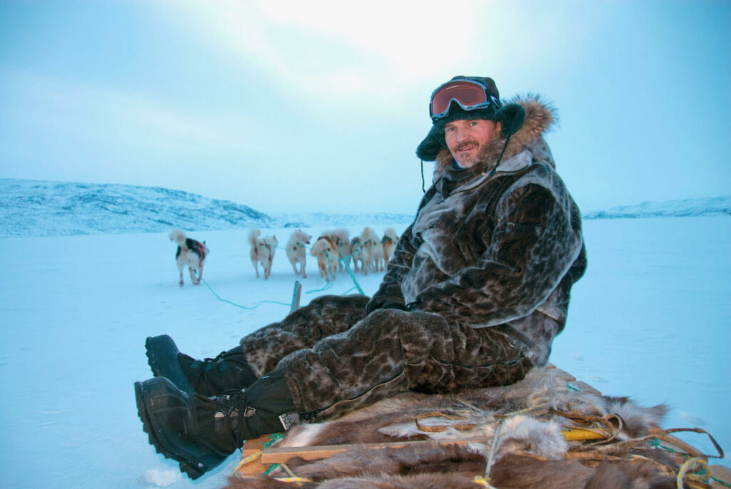 Photo of Kieran Meeke riding a sled harnessed to a pack of dogs in Greeland